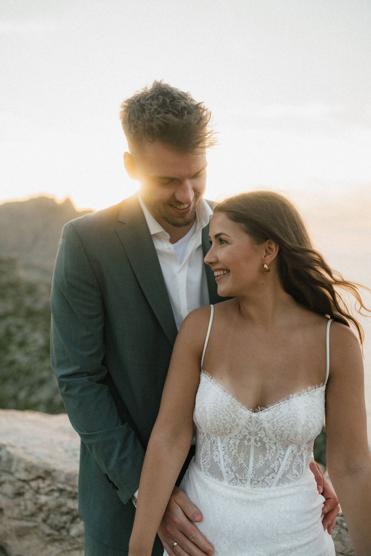 Smiling bride in lace gown gazes at groom in gray suit during golden sunset outdoor wedding shoot.