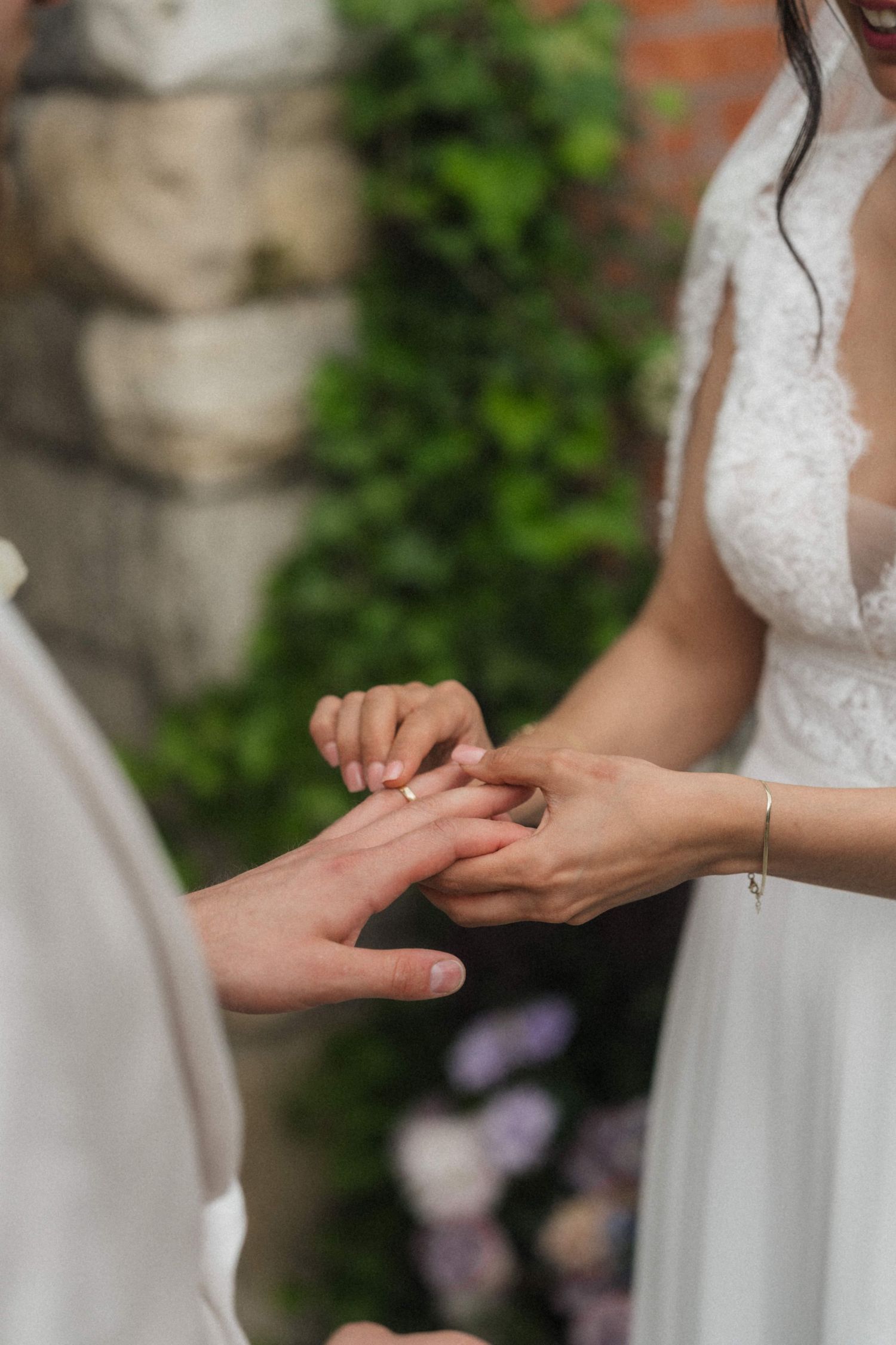 Close-up of bride placing wedding ring on grooms hand during outdoor ceremony with floral backdrop.