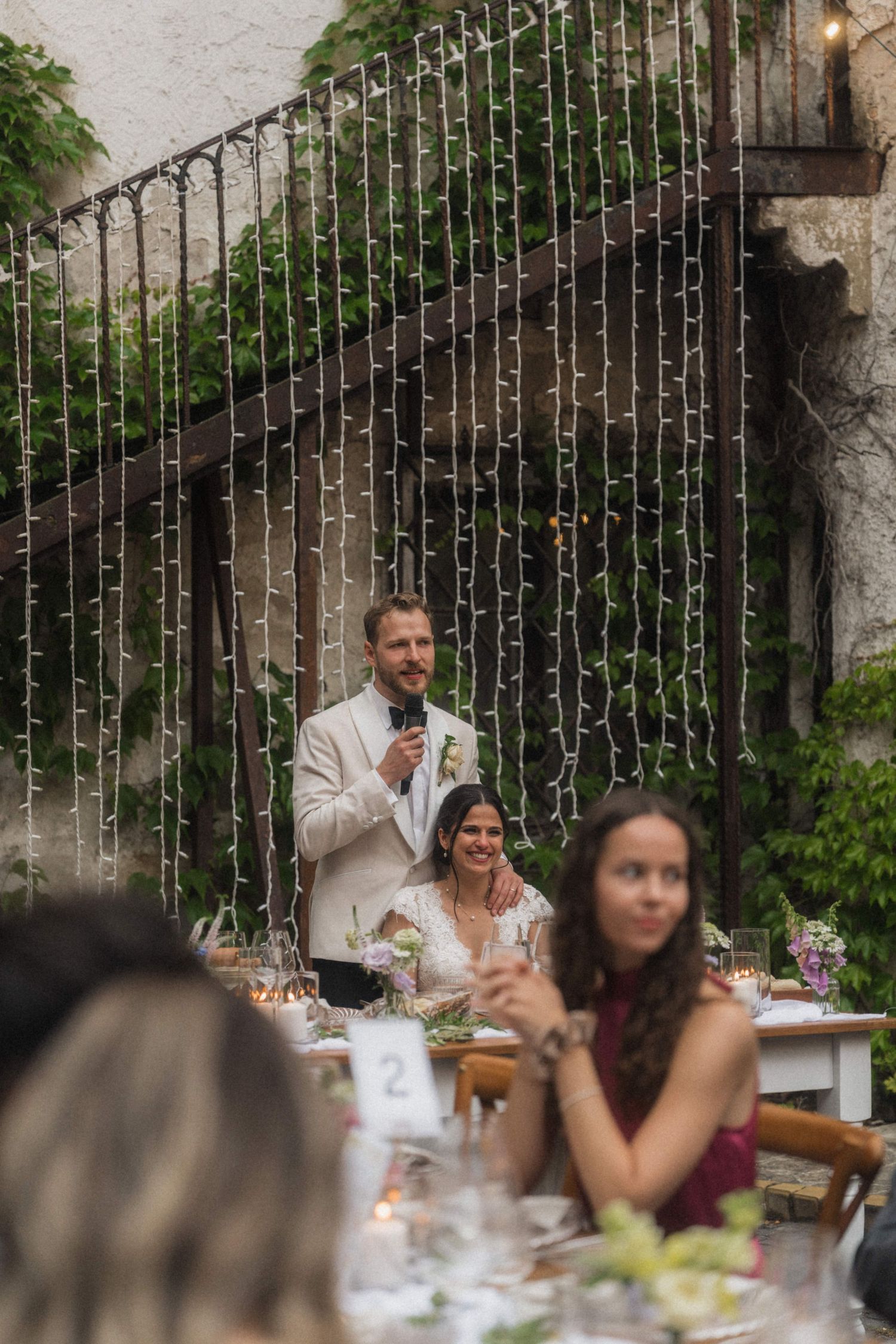Groom gives speech at outdoor wedding reception with fairy light backdrop while bride sits smiling nearby.