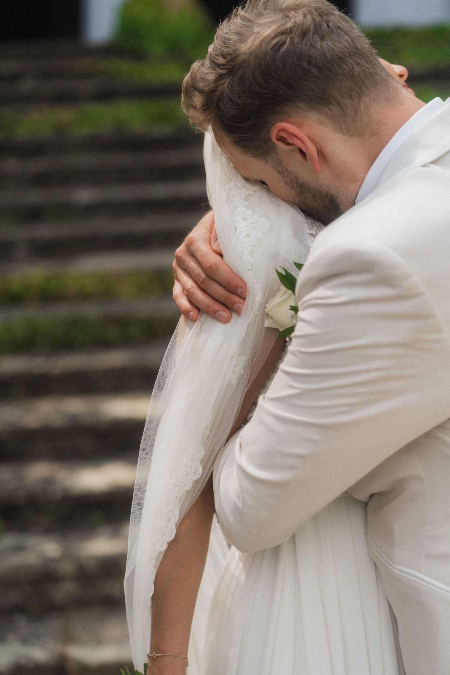 Groom in white suit embracing bride in lace wedding dress and veil outdoors near stone steps.