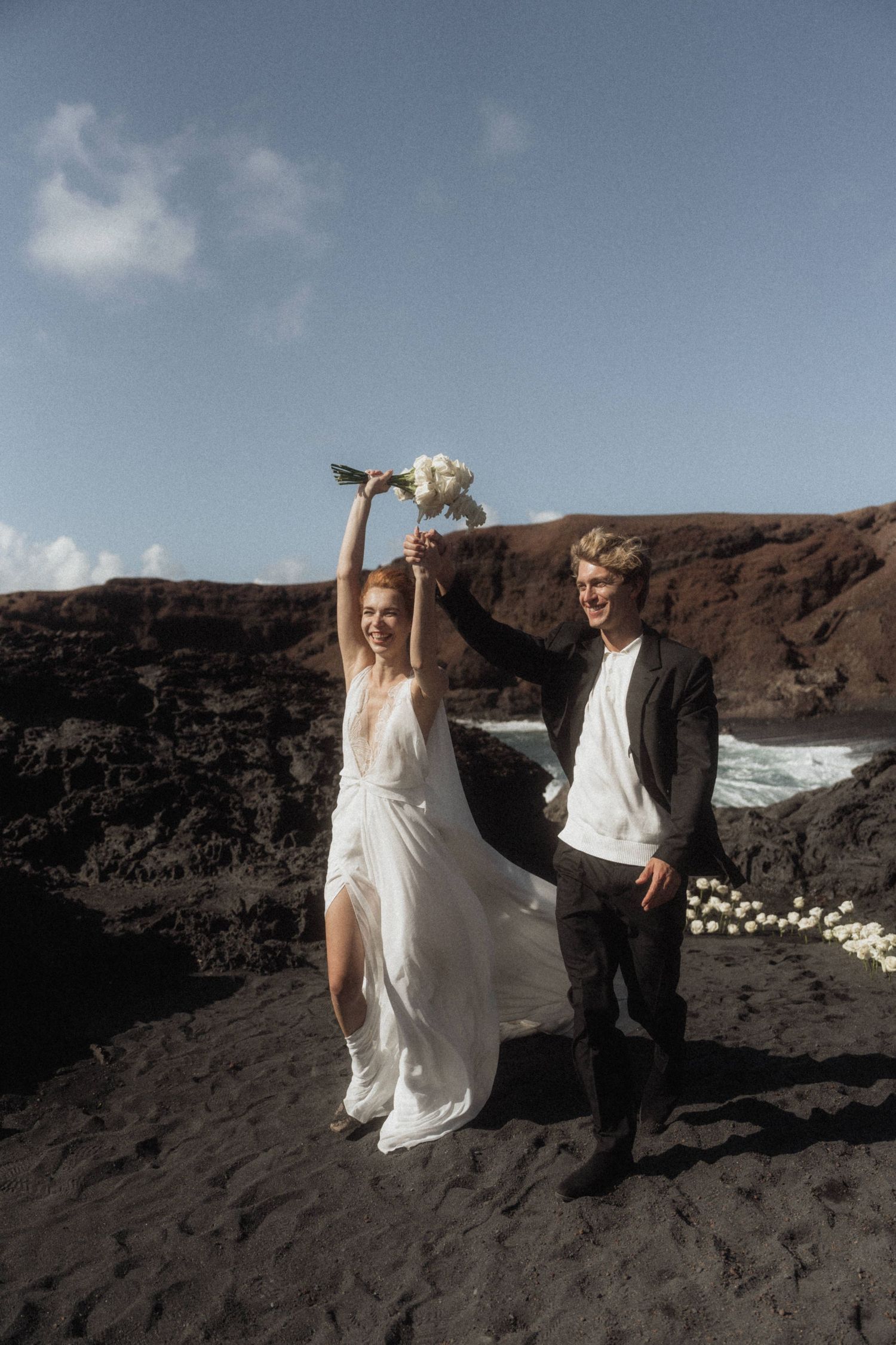 Bride and groom celebrating on black volcanic beach holding bouquet aloft under blue sky.