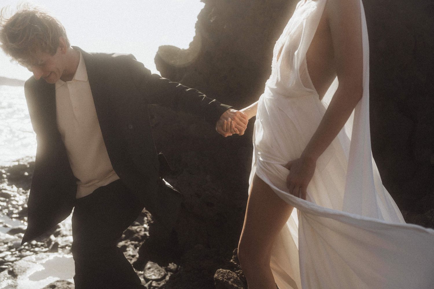 Bride and groom hold hands navigating rocky coastal terrain in wedding attire.