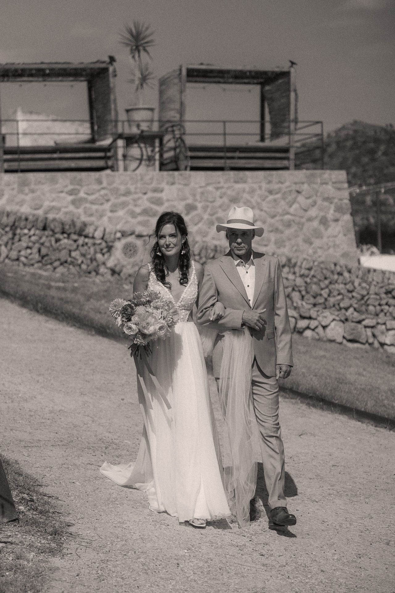 Bride and groom walk together on rustic path, bride holding bouquet, groom wearing hat, in black and white.