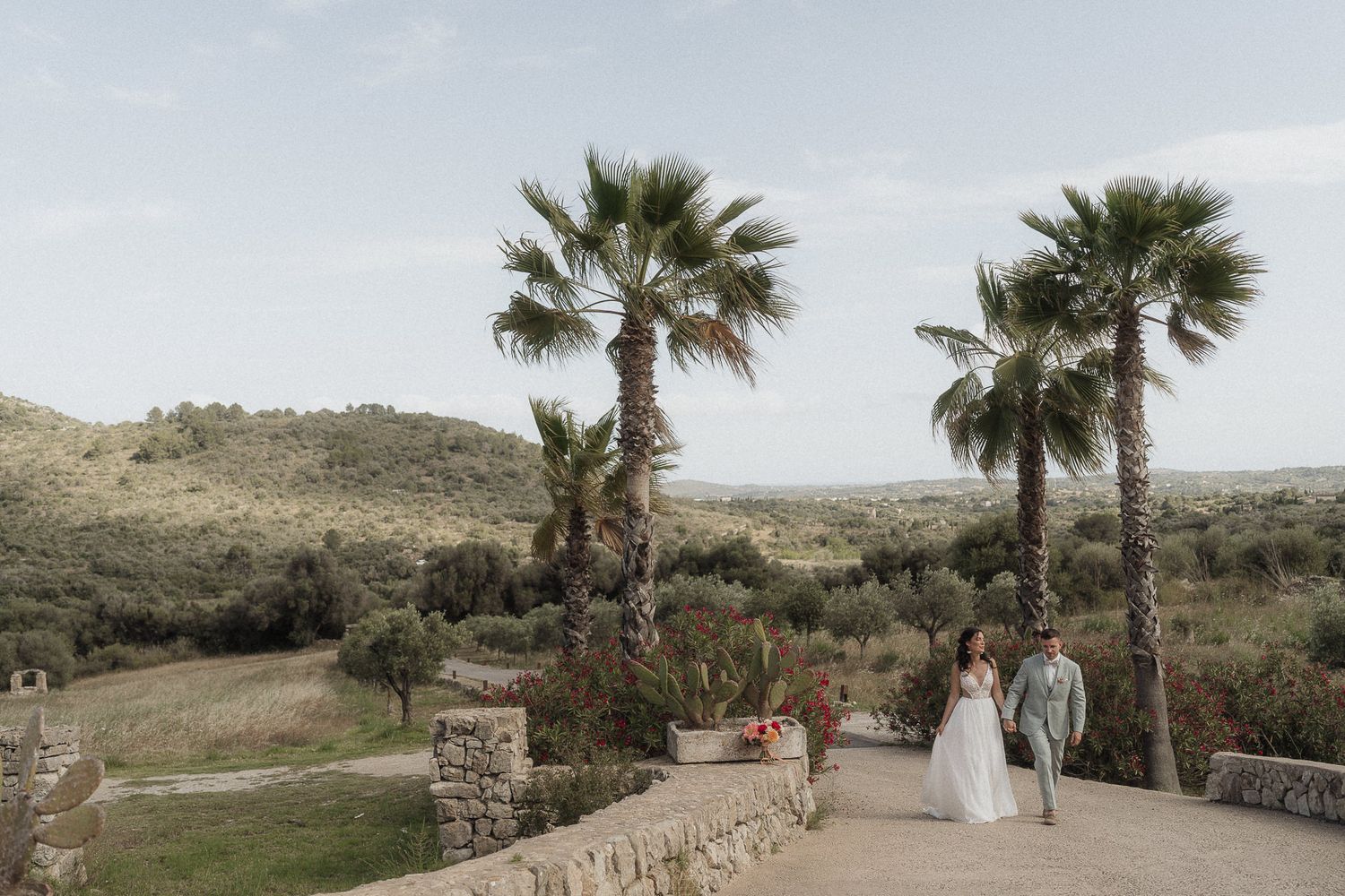 Bride and groom walk hand in hand on a stone terrace with palm trees and rolling green hills at a scenic outdoor wedding.