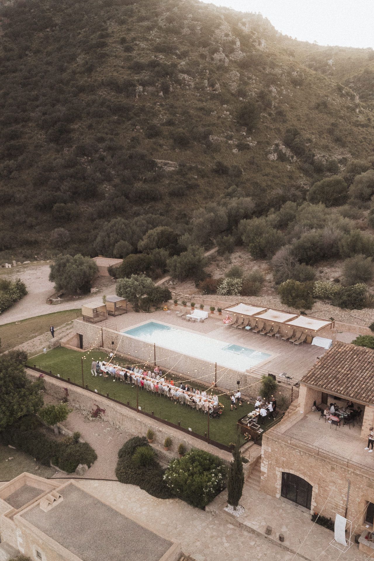 Aerial view of a rustic Mediterranean villa with a swimming pool and outdoor wedding reception, nestled against a lush hillside.