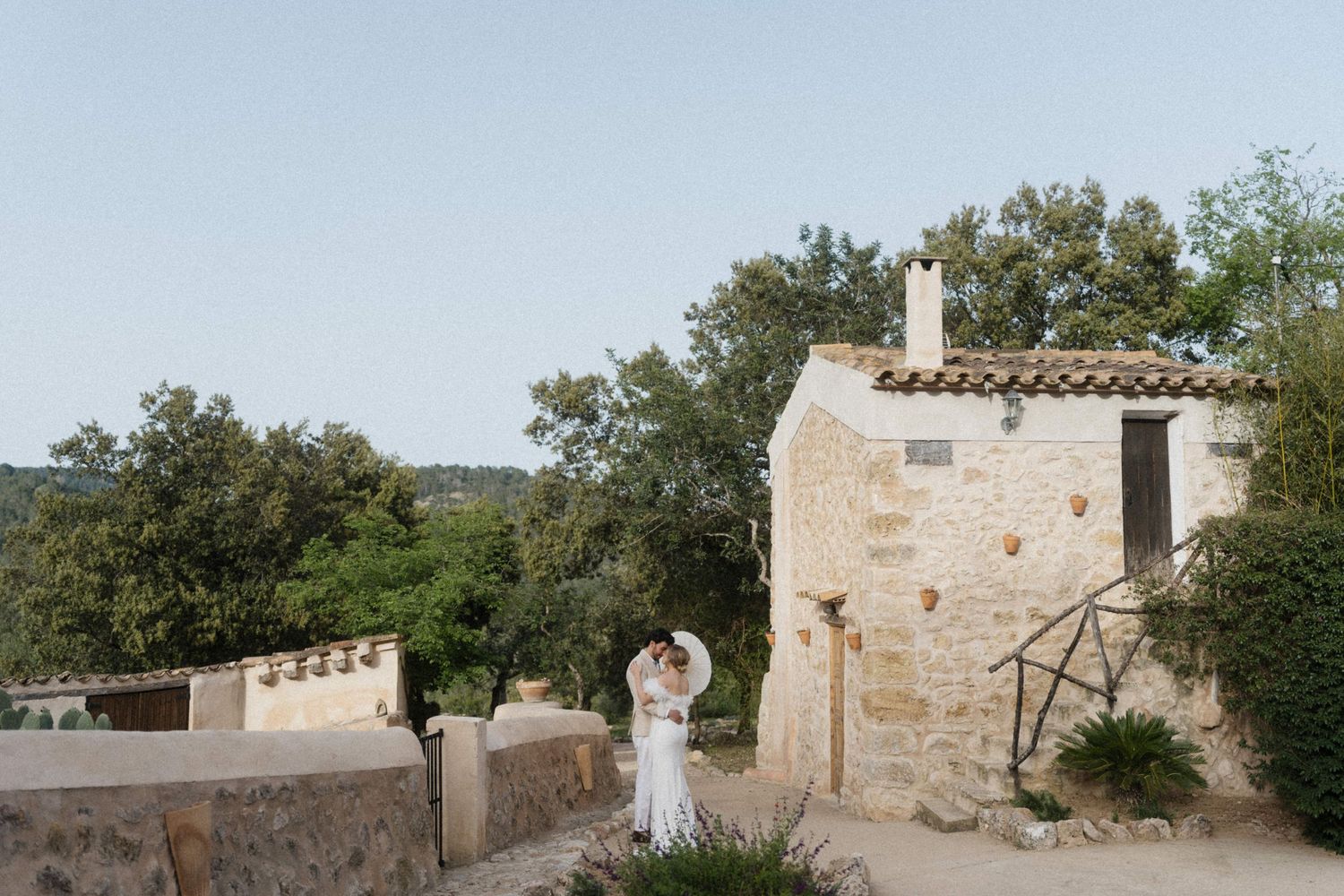 Couple embraces near a rustic stone building surrounded by lush trees at a wedding venue.