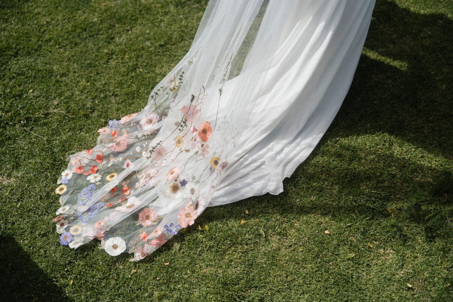 Close-up of a bridal veil train adorned with delicate colorful floral embroidery resting on green grass.