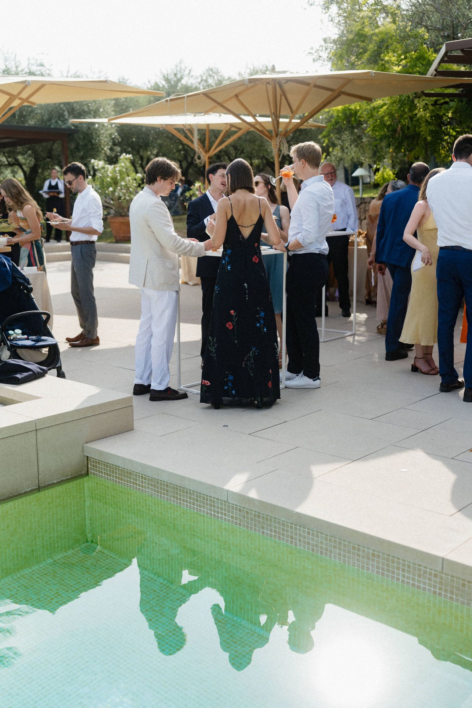 Elegant outdoor wedding reception guests mingling poolside under large umbrellas at a sunny venue.