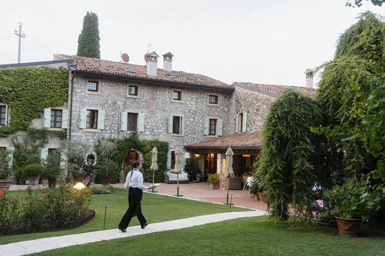 A person walks through a lush garden courtyard of a rustic stone Italian villa surrounded by greenery.