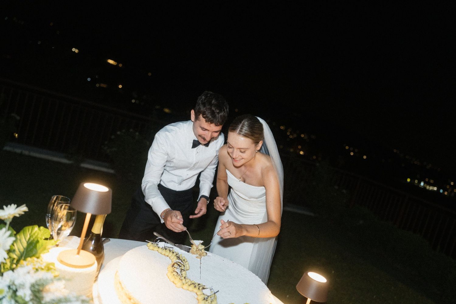 Bride and groom cutting their wedding cake together outdoors at night, illuminated by warm ambient lighting.