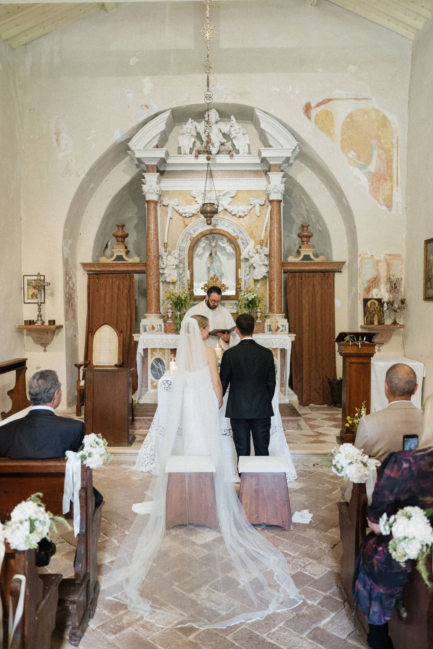 Bride and groom exchange vows at the altar of a charming Italian chapel with frescoed walls and white floral decor.