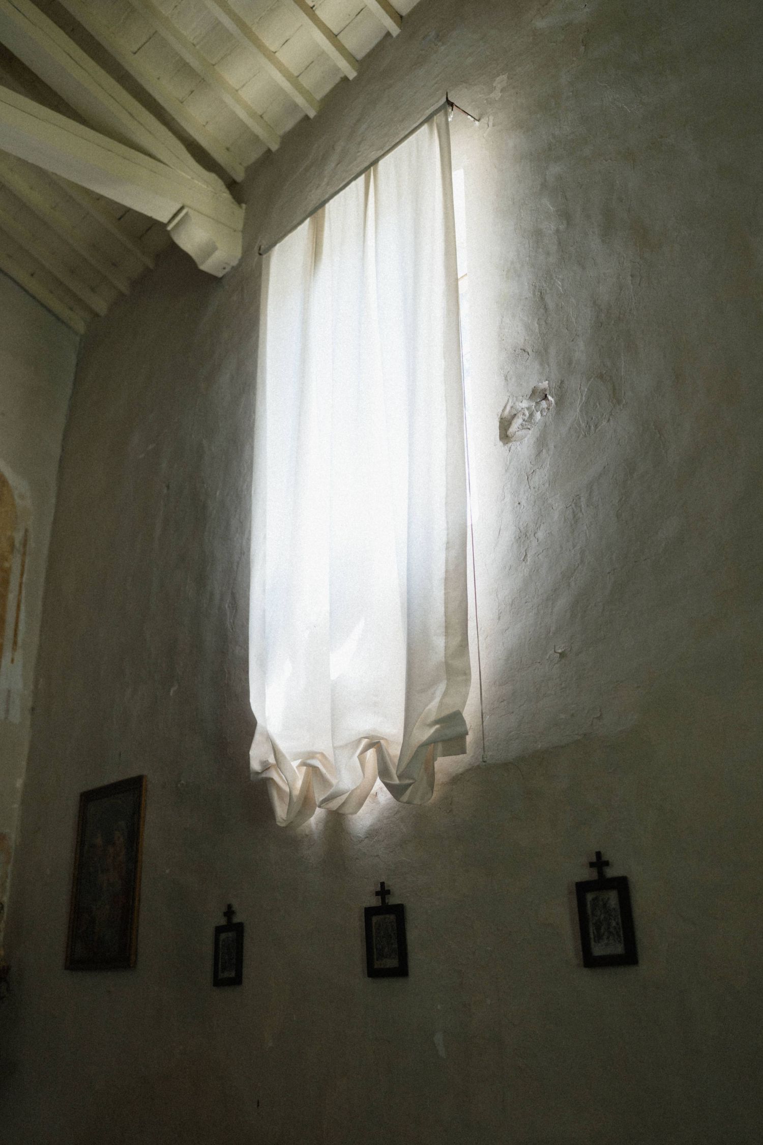 Rustic stone chapel interior with tall narrow window, white curtain, and small religious framed pictures on wall.