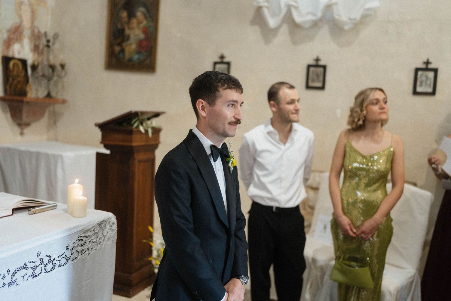 Groom in black tuxedo with yellow boutonniere stands at church altar with guests in elegant attire behind him.