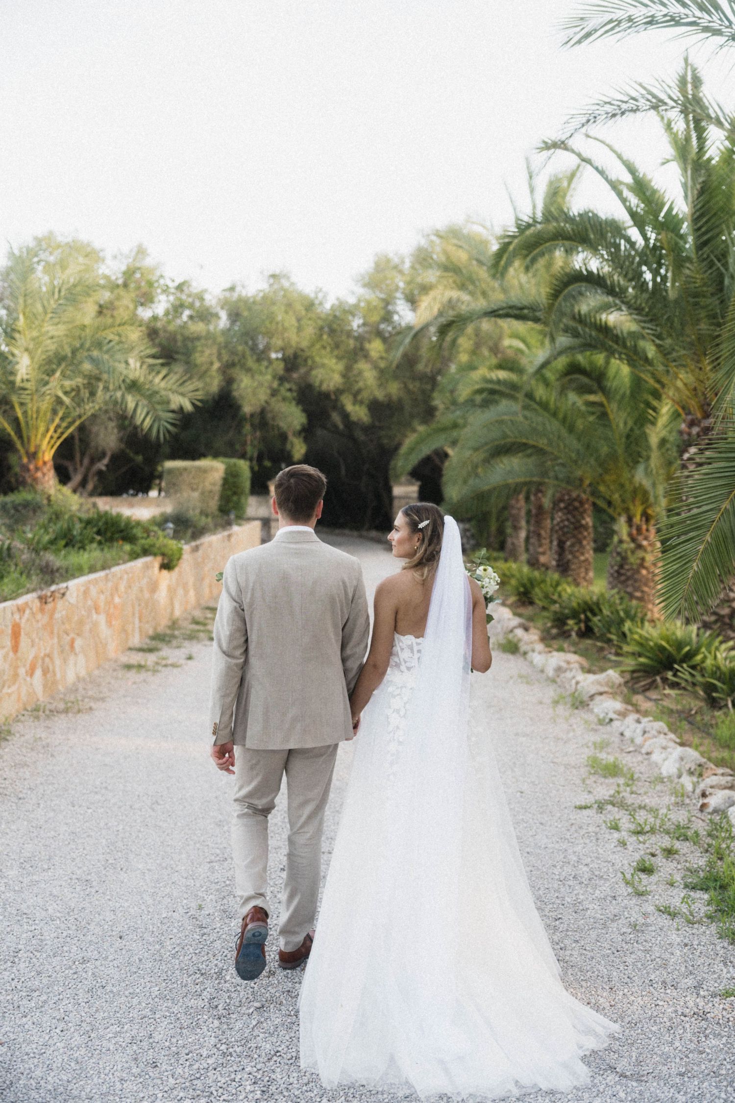 Bride in white gown and groom in tan suit walking hand in hand along a palm tree-lined path.