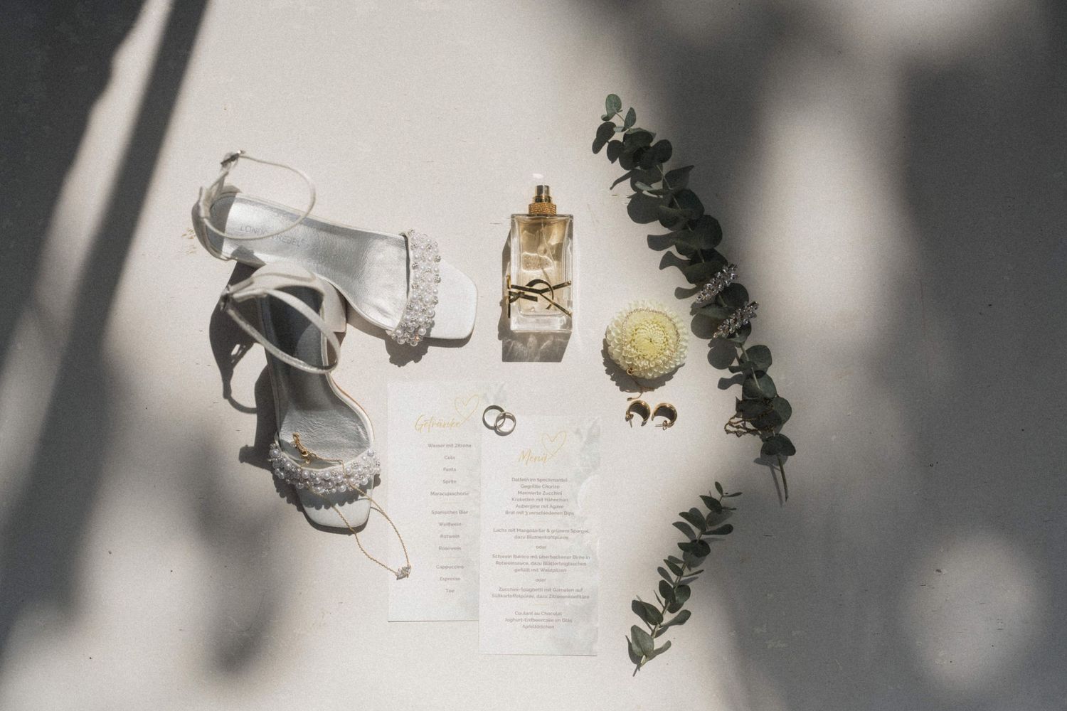 Elegant wedding flat lay featuring white heeled sandals, perfume, rings, and eucalyptus on white surface.