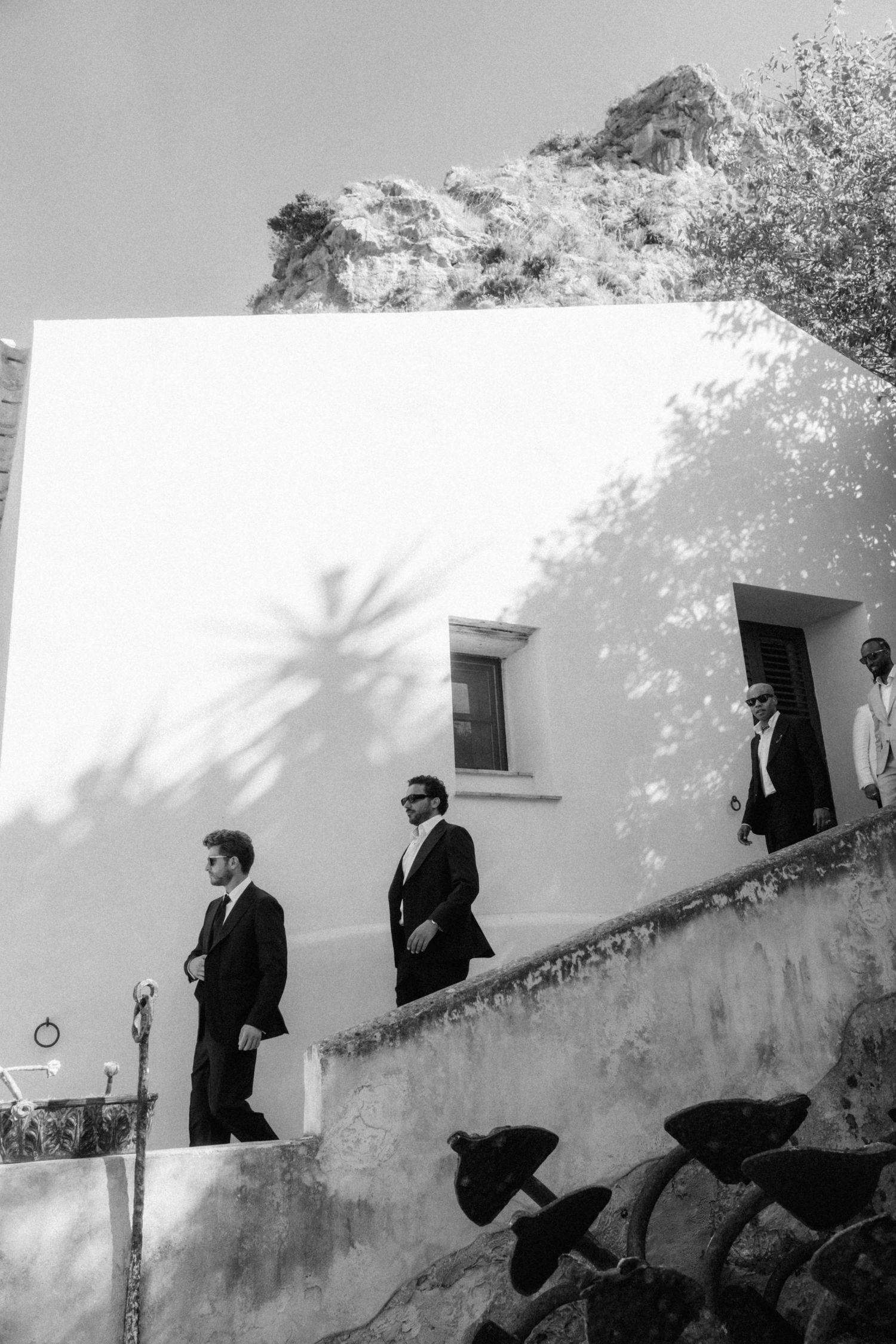 Black and white photo of groom and groomsmen in dark suits descending stone steps at Mediterranean wedding venue.
