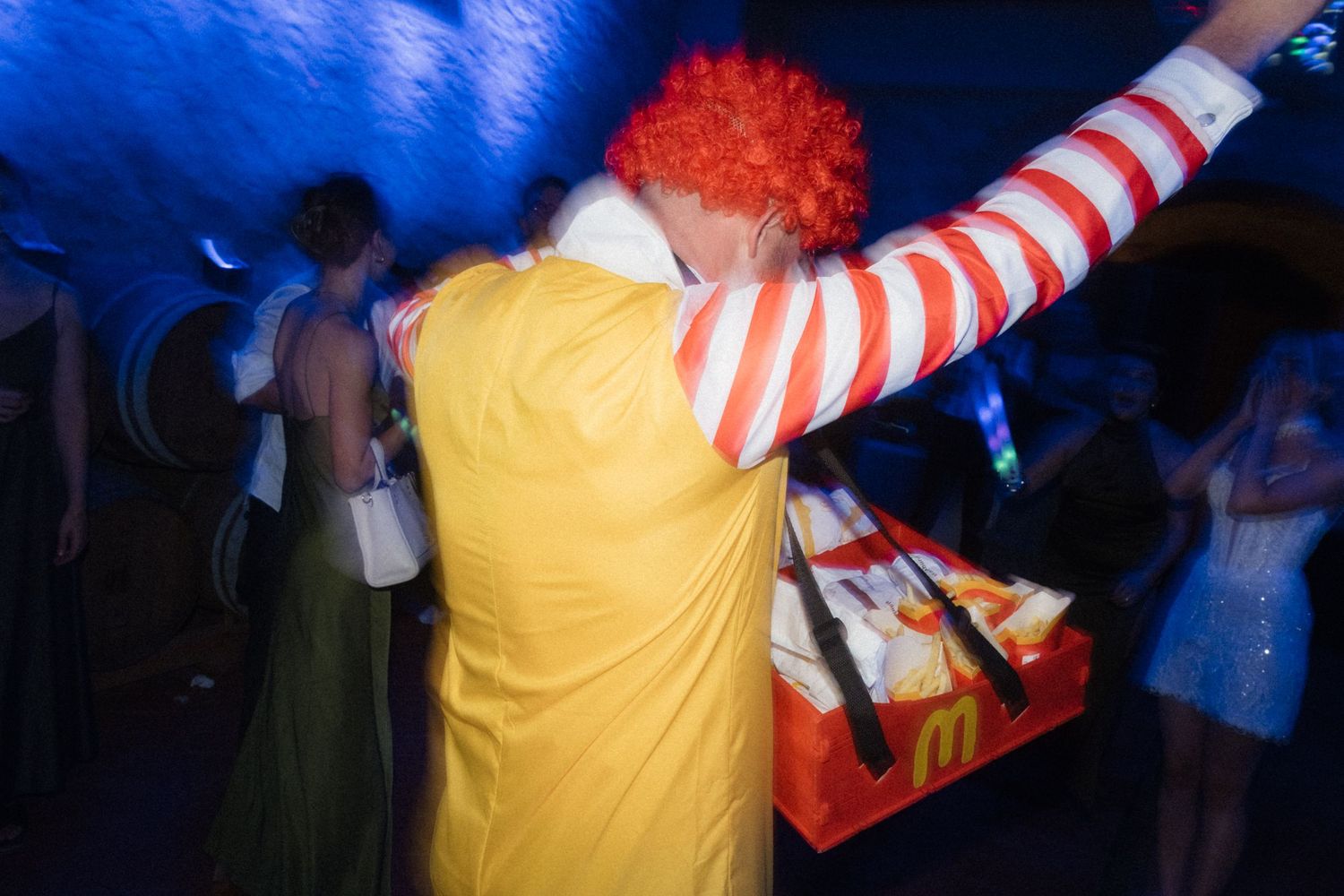 Person dressed as Ronald McDonald dancing at a lively wedding reception party with blue lighting.