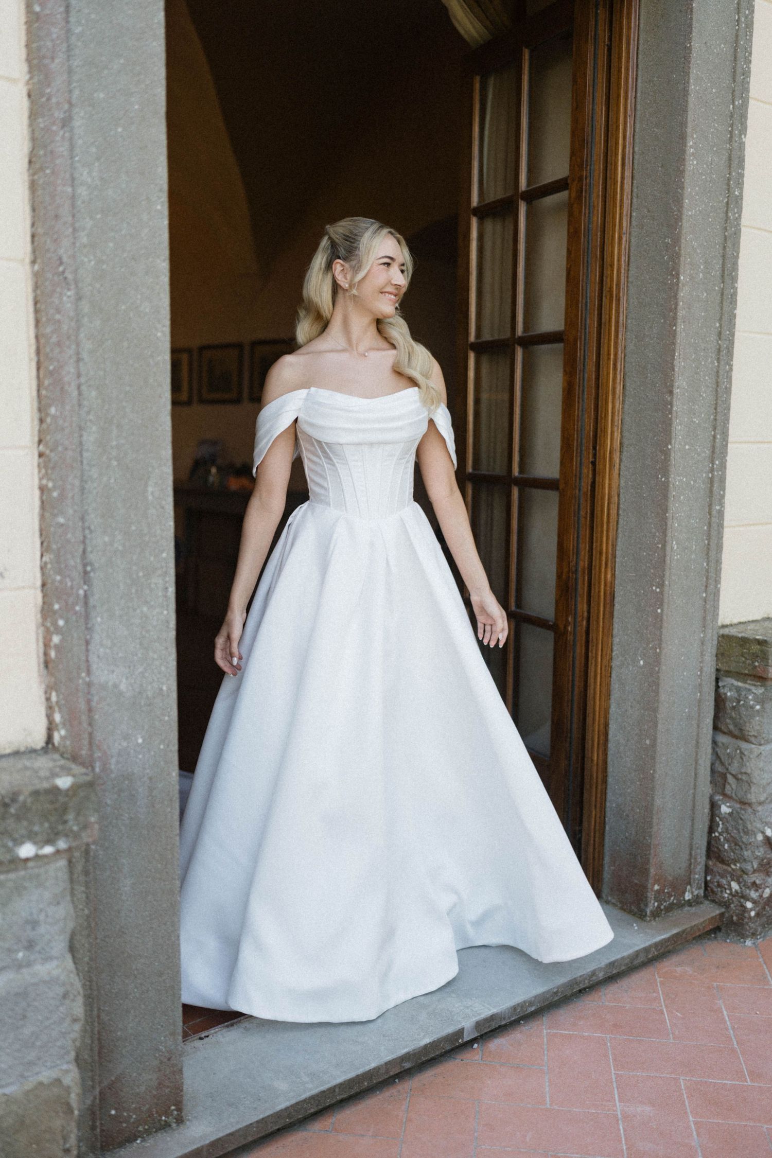 Smiling bride in elegant off-shoulder white ball gown standing in a stone doorway.