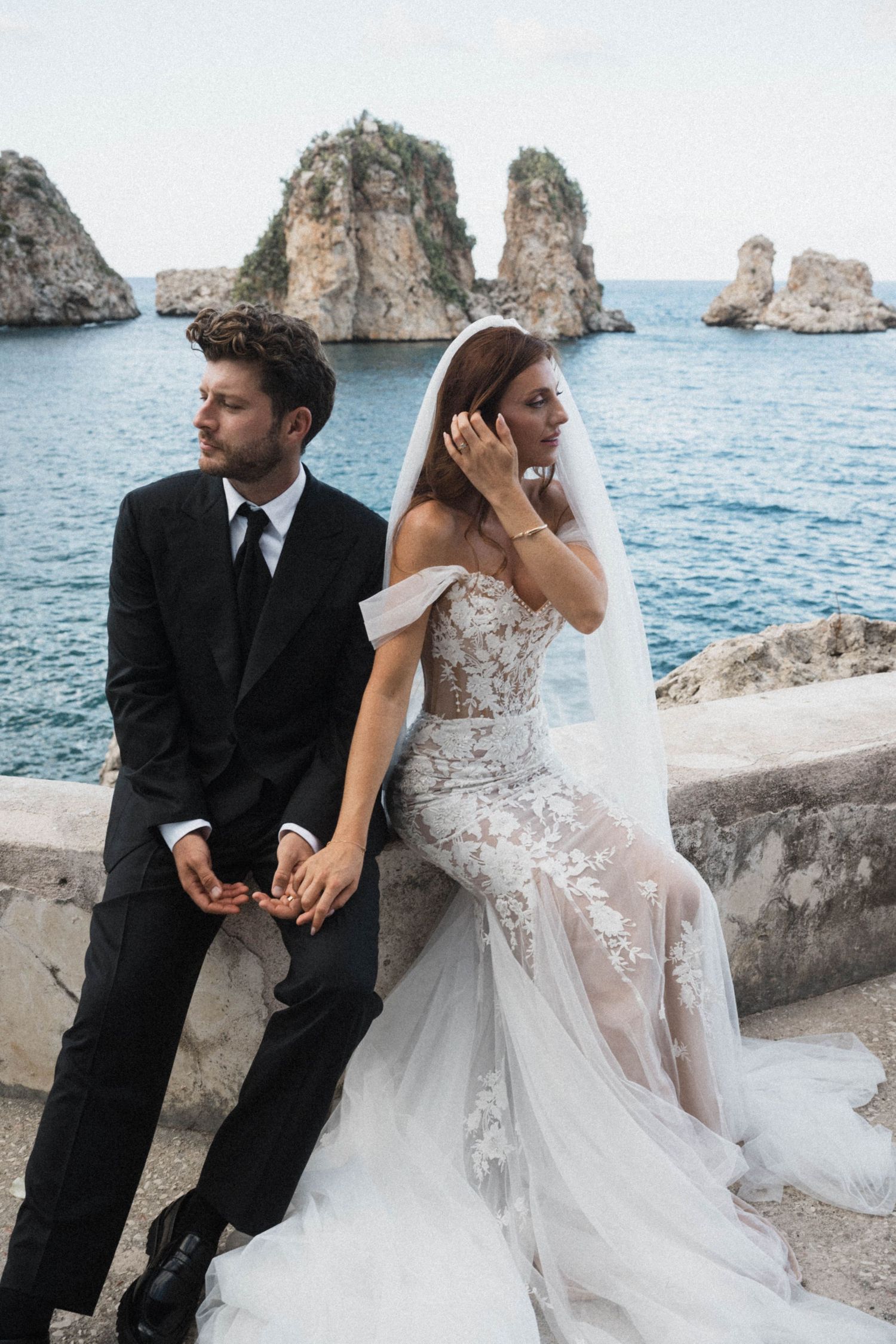 Bride and groom sitting on stone wall with dramatic sea stacks and blue ocean backdrop at coastal wedding.