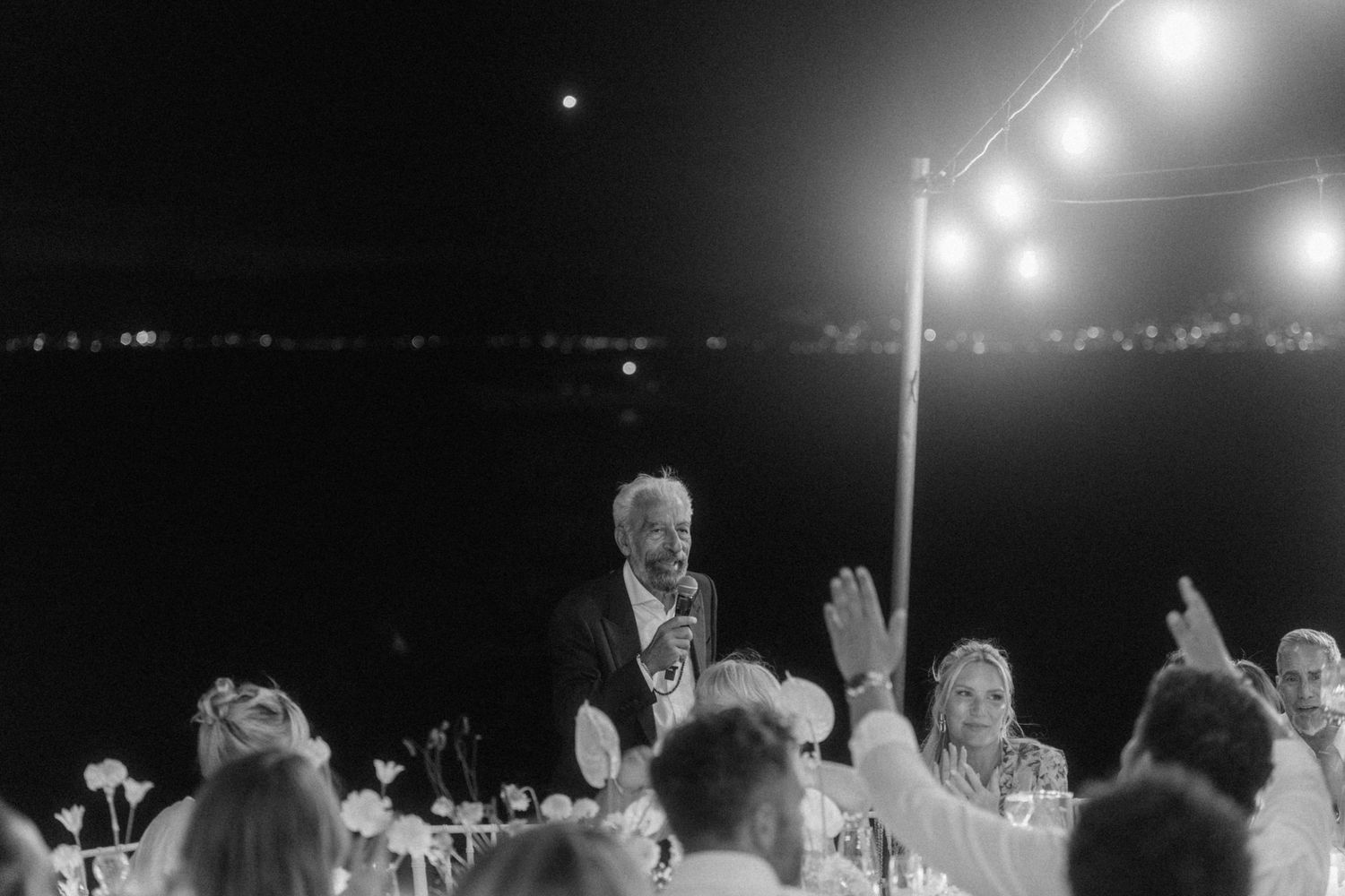 Black and white photo of a man giving a toast speech at an outdoor wedding reception under string lights.