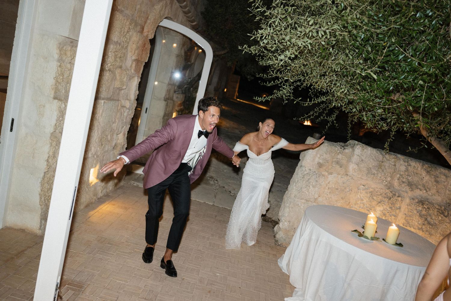 A groom in a burgundy blazer and a bride in a white gown dance joyfully near a candlelit table at a night wedding.