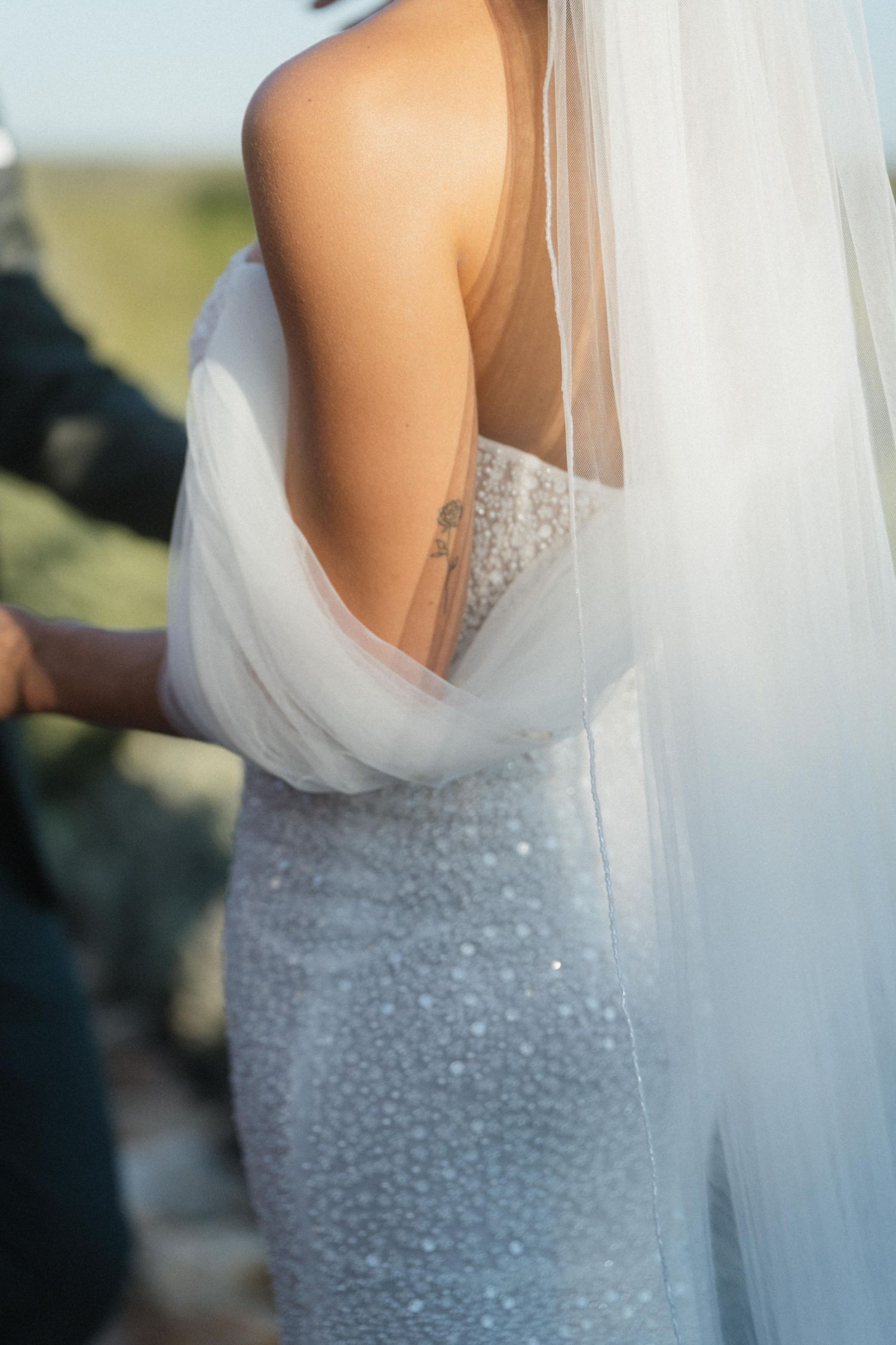 Close-up of a bride's back showing a sparkling beaded wedding gown with sheer off-shoulder drape and a long flowing tulle veil.