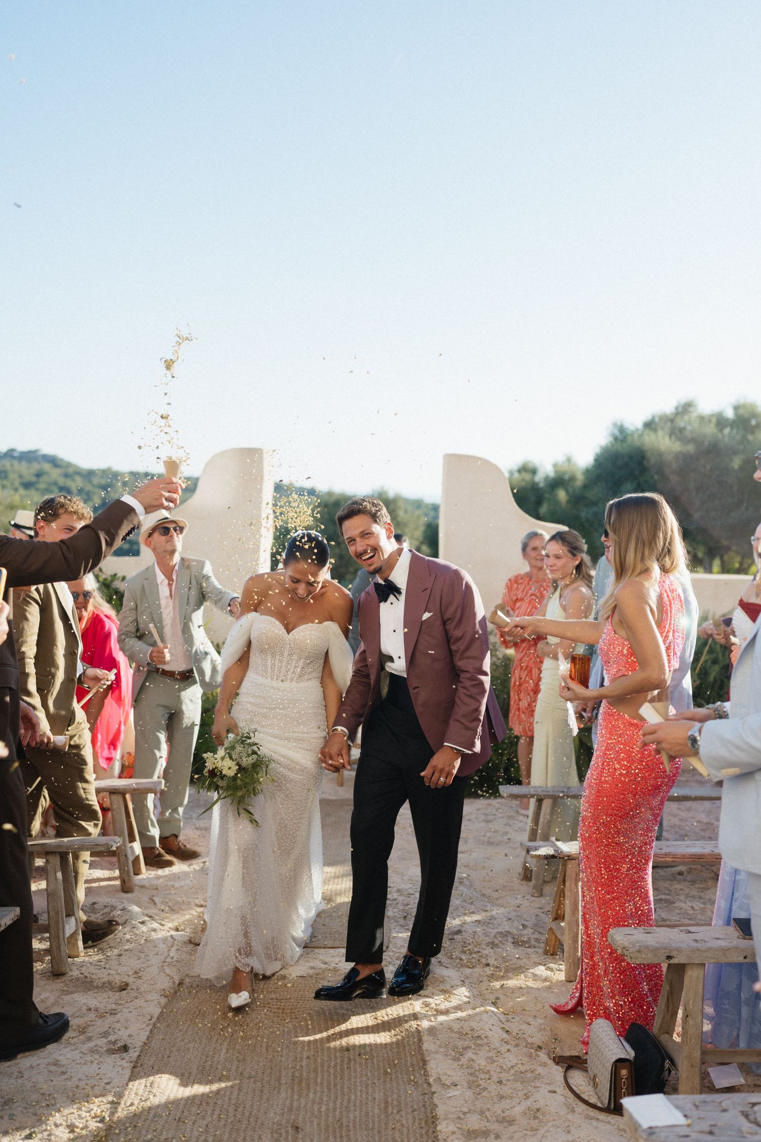 Smiling newlyweds walk hand in hand down the aisle as guests cheer and toss confetti at an outdoor wedding.