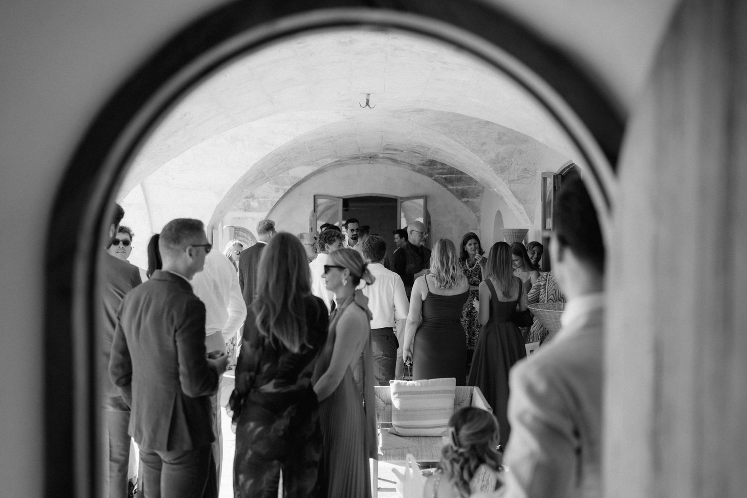 Black and white photo of wedding guests mingling and chatting beneath a rustic stone archway at a celebration.