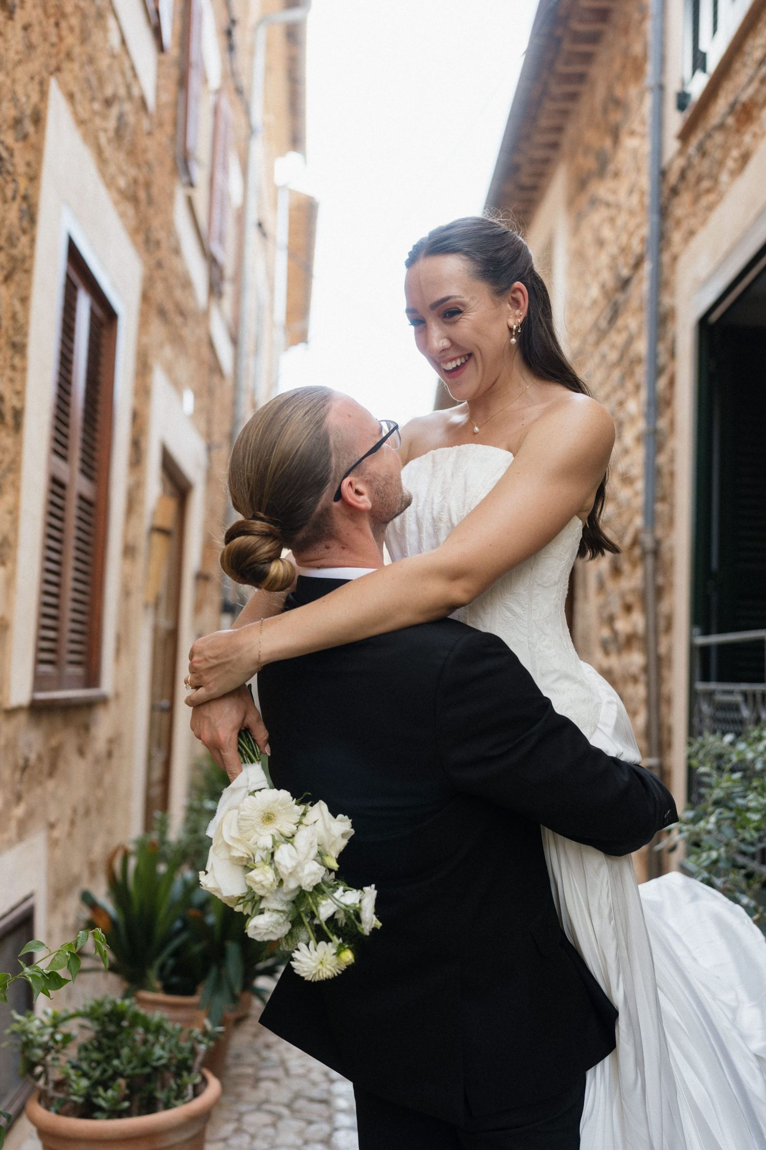 Smiling bride in a white dress is lifted by her groom, who holds a white bouquet, in a stone village alley.