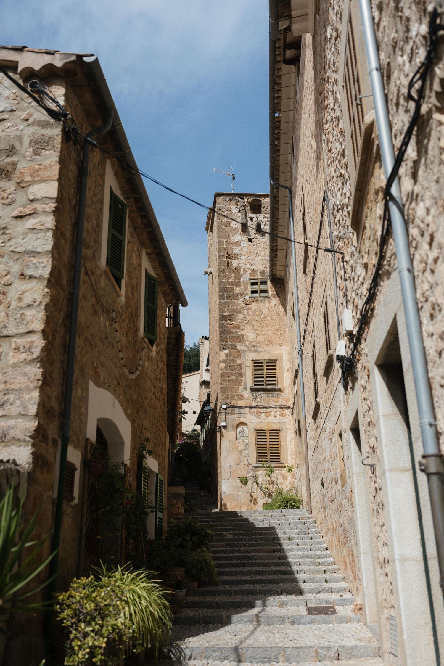 Narrow stone staircase winding between rustic medieval buildings with green shutters and a weathered bell tower in a Mediterranean village.