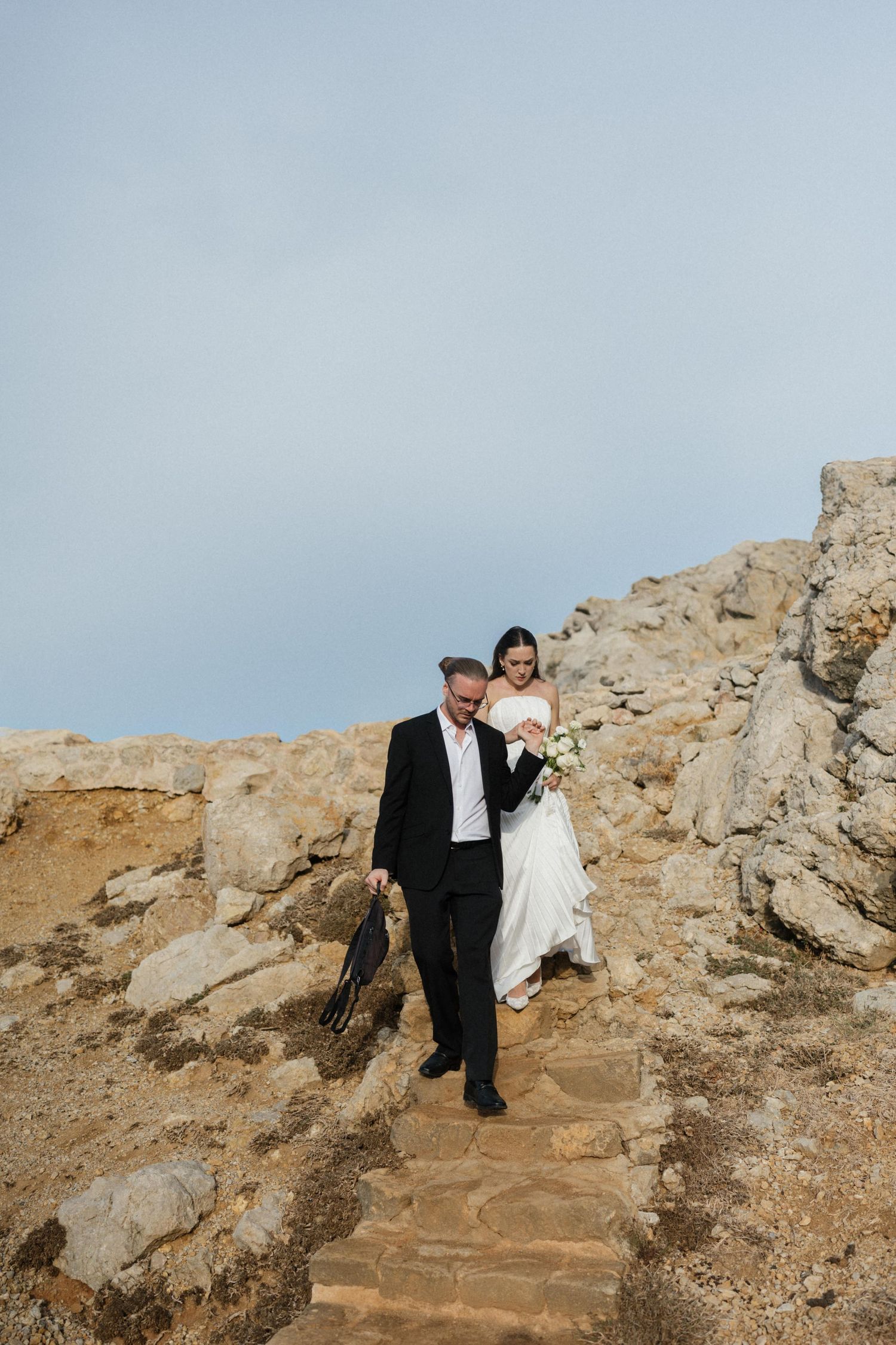 A bride in a white dress holding a bouquet walks down rustic stone steps beside a groom in a black suit on a rocky hillside.