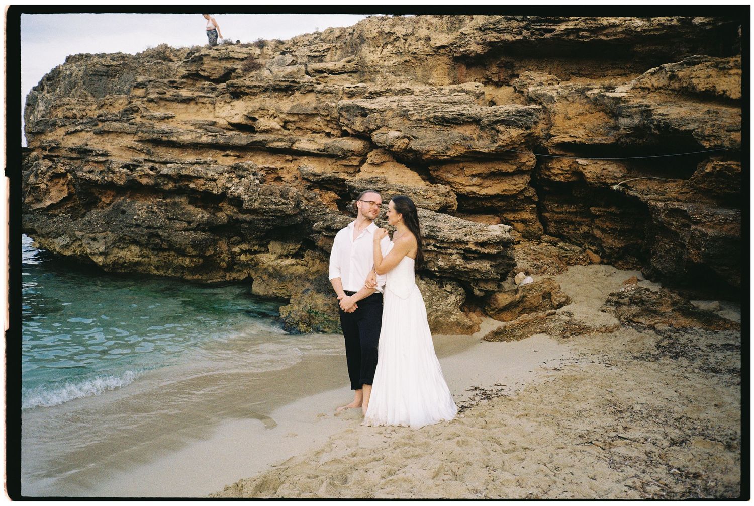 Bride in a white gown and groom in a white shirt embrace on a sandy beach beside turquoise water and rugged limestone cliffs.