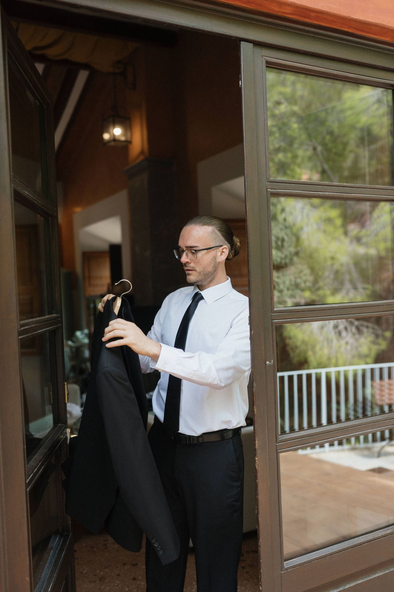 Man in glasses, white dress shirt and black tie holds a suit jacket on a hanger in a rustic doorway with glass doors and a view of trees.