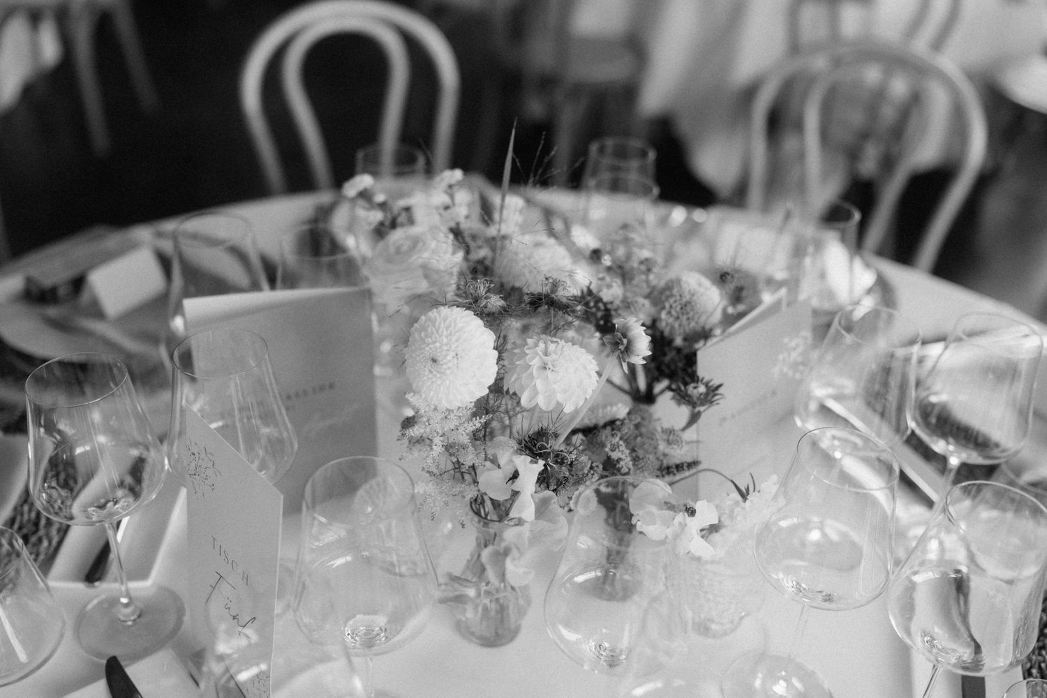 Black and white photo of an elegant round wedding reception table with floral centerpiece and crystal glassware.