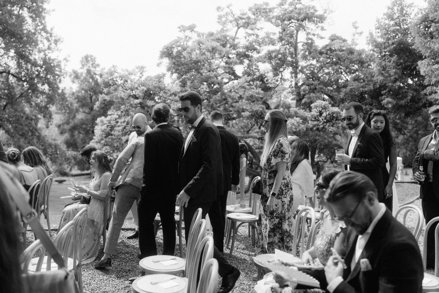 Black and white outdoor wedding reception with elegantly dressed guests mingling among chairs and lush trees.