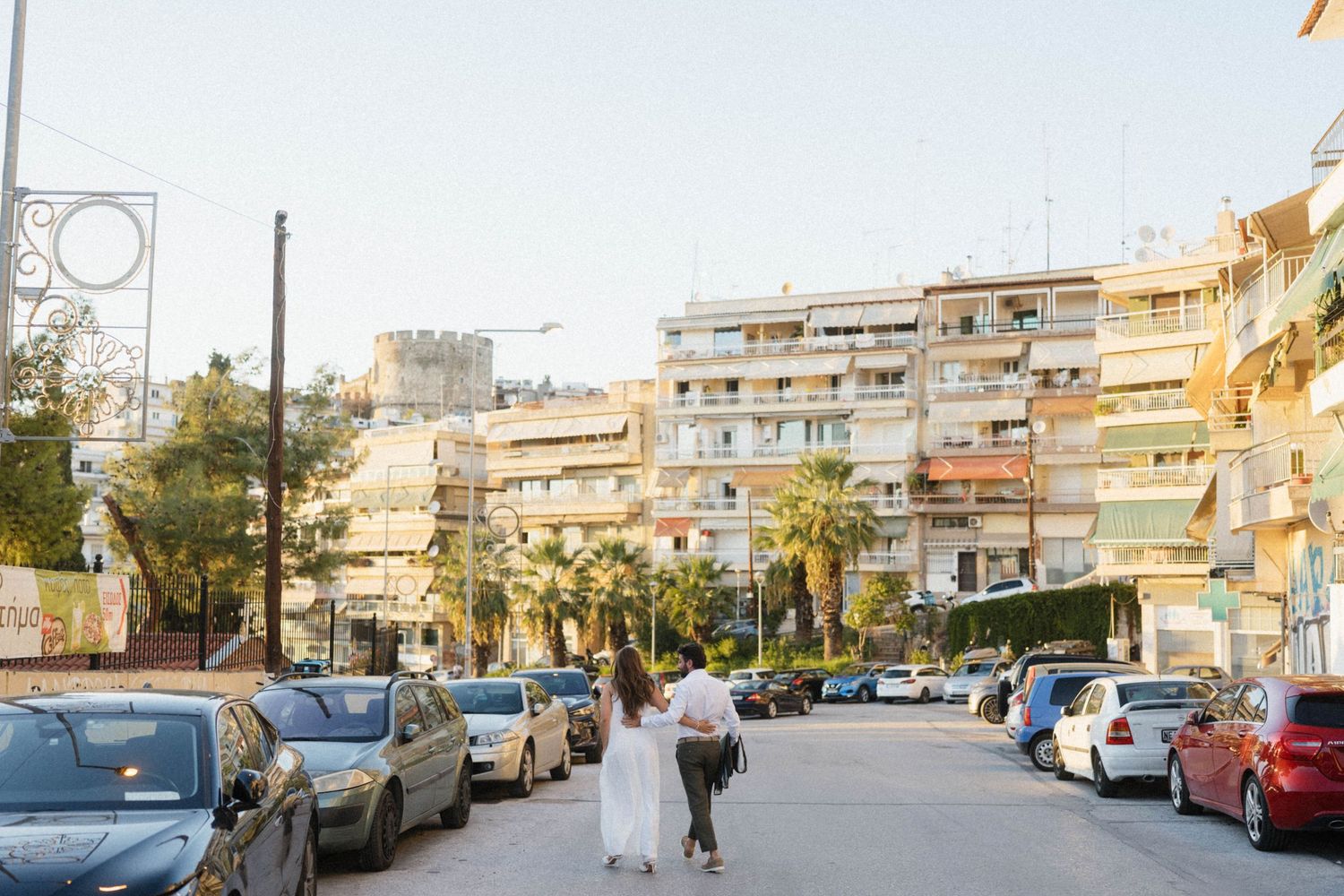 Couple walking hand-in-hand through a sunny Mediterranean street lined with parked cars and palm trees.