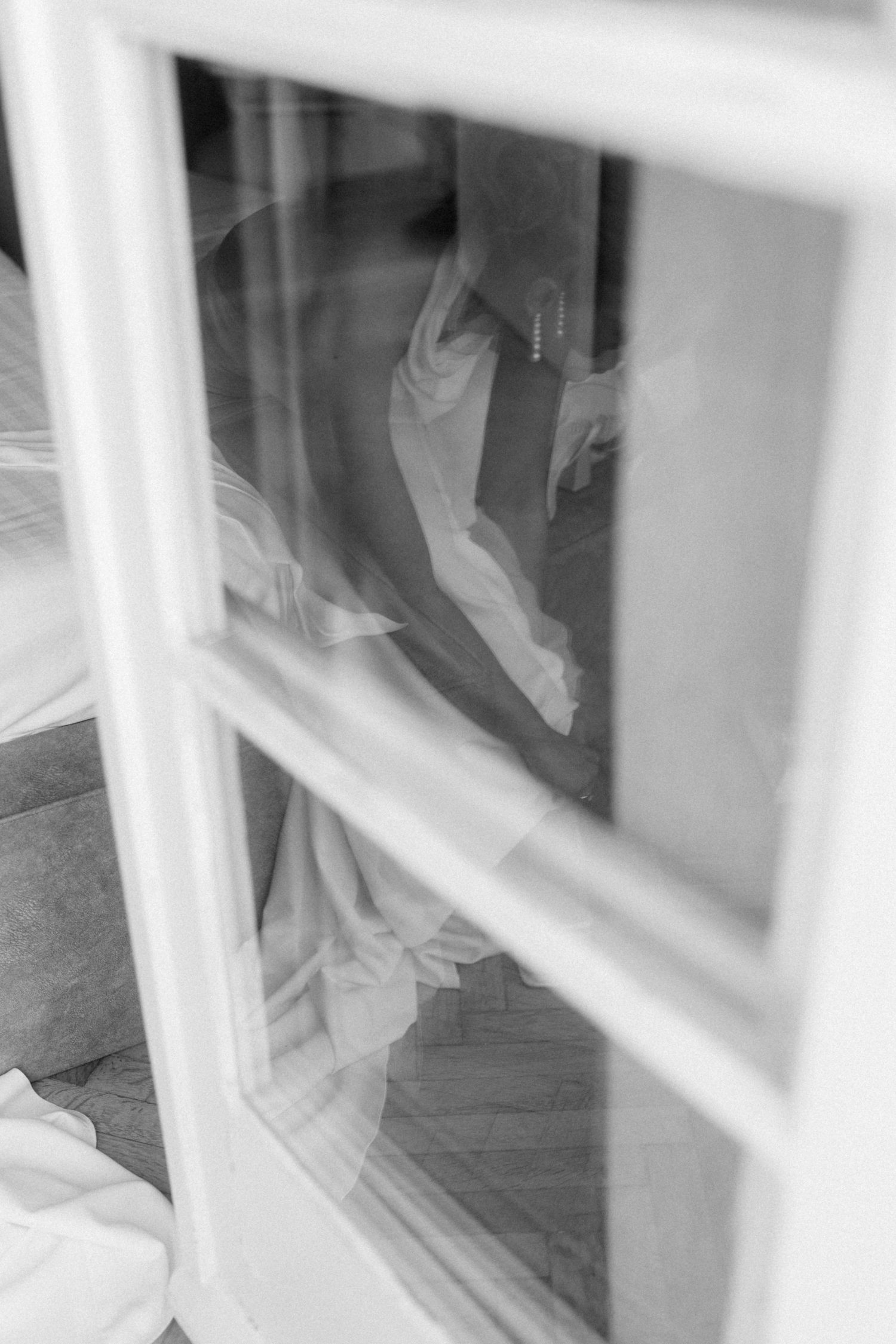 Black and white reflection of bride in wedding dress seen through window panes.