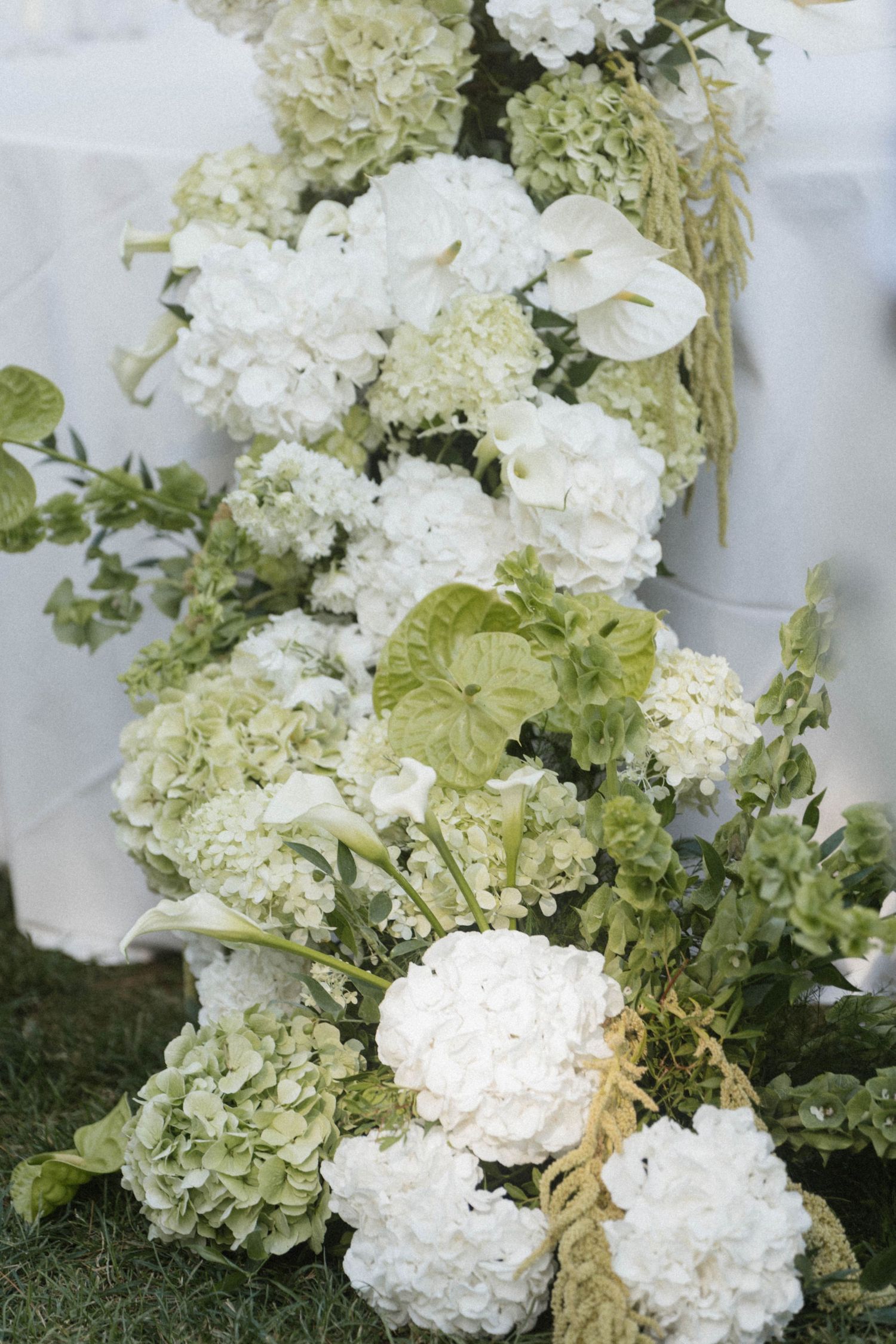 Elegant white hydrangea and green anthurium floral arrangement cascading along a table at an outdoor wedding reception.