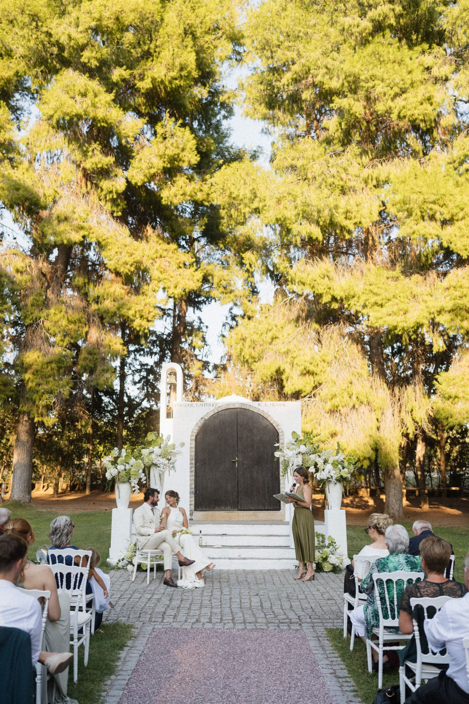 Outdoor wedding ceremony with couple seated at altar before rustic wooden doors, surrounded by tall trees and white floral arrangements.