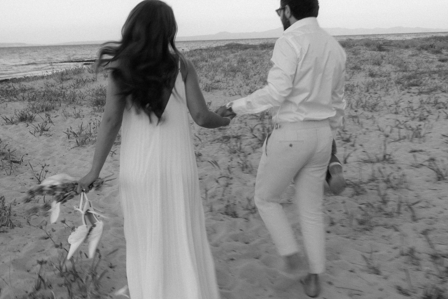 Black and white photo of a couple in white outfits holding hands walking on a sandy beach.