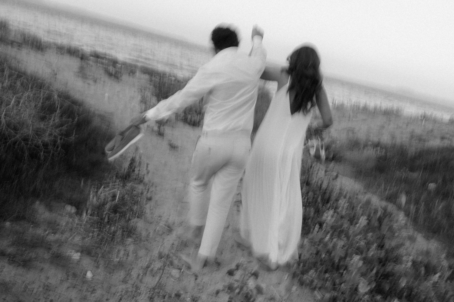 Black and white photo of a couple in white attire walking hand in hand through coastal dunes.