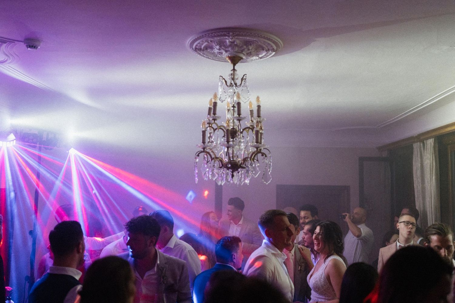 Wedding reception dance floor filled with guests dancing under colorful laser lights and a crystal chandelier.