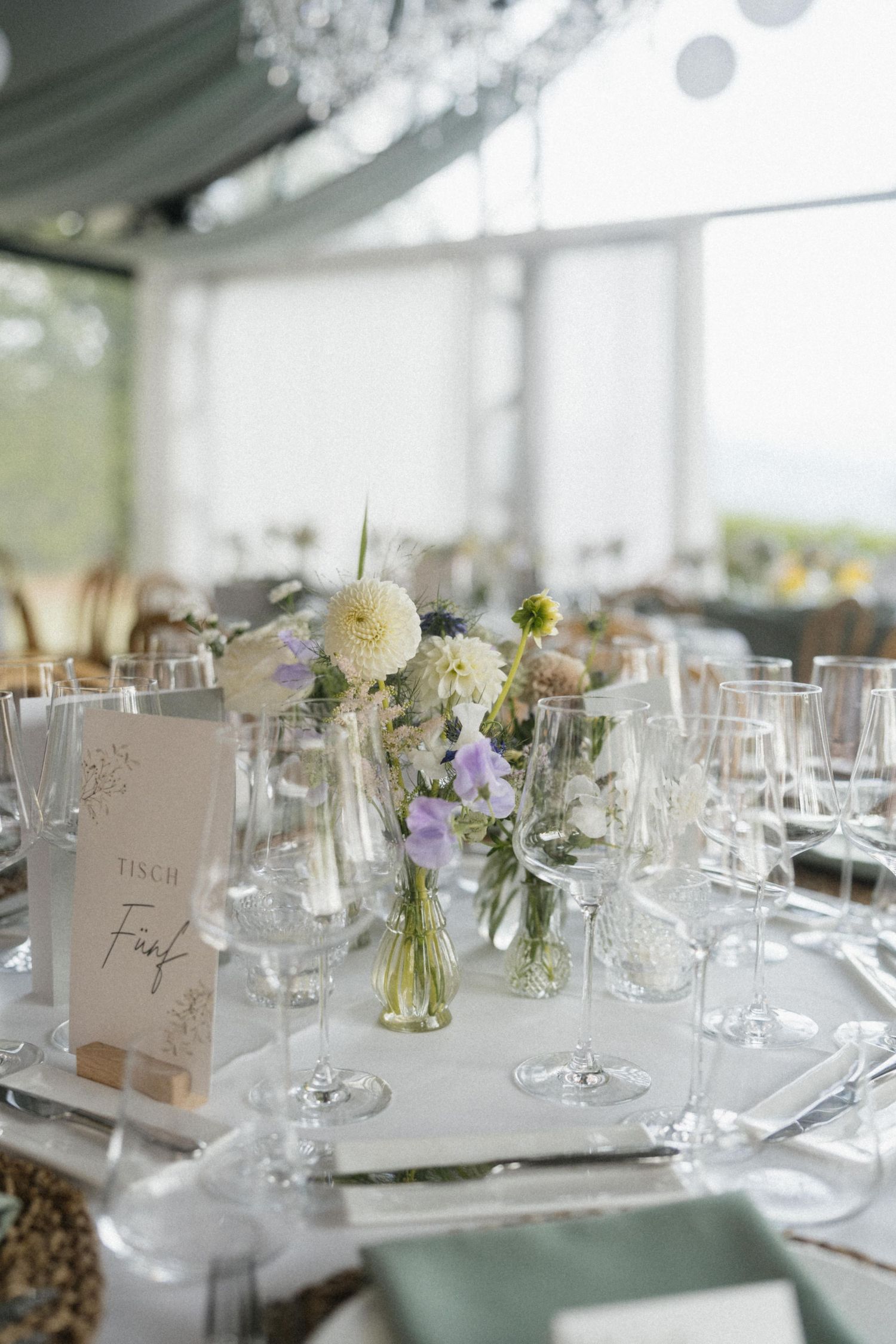 Elegant wedding reception table set with crystal glasses, white and purple floral centerpiece, and a handwritten table number card.