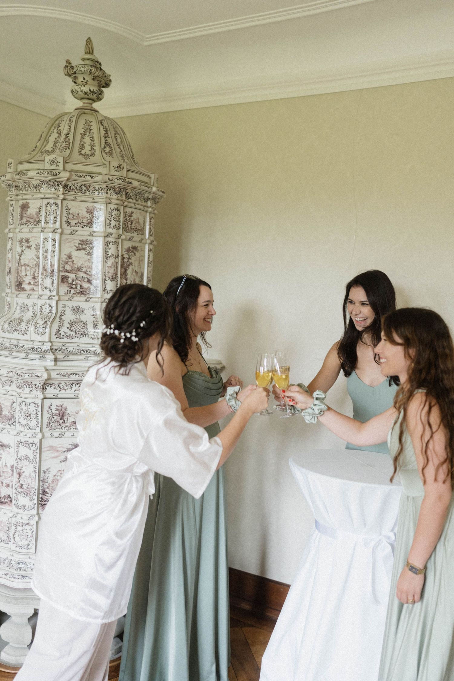 Bridesmaids in sage green dresses toasting champagne with bride in white robe near ornate fireplace.