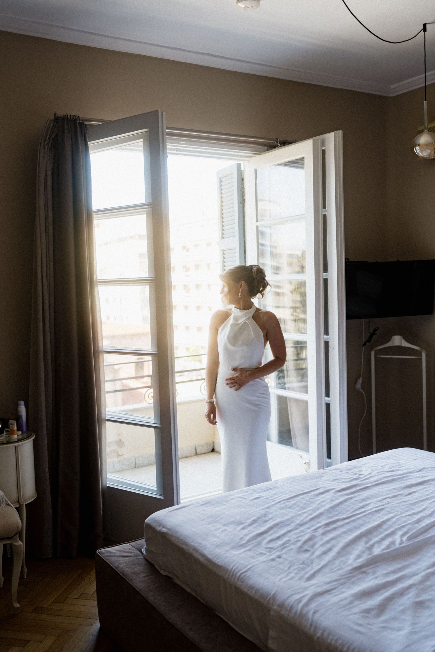 Bride in white halter gown stands by open French doors in sunlit bedroom.