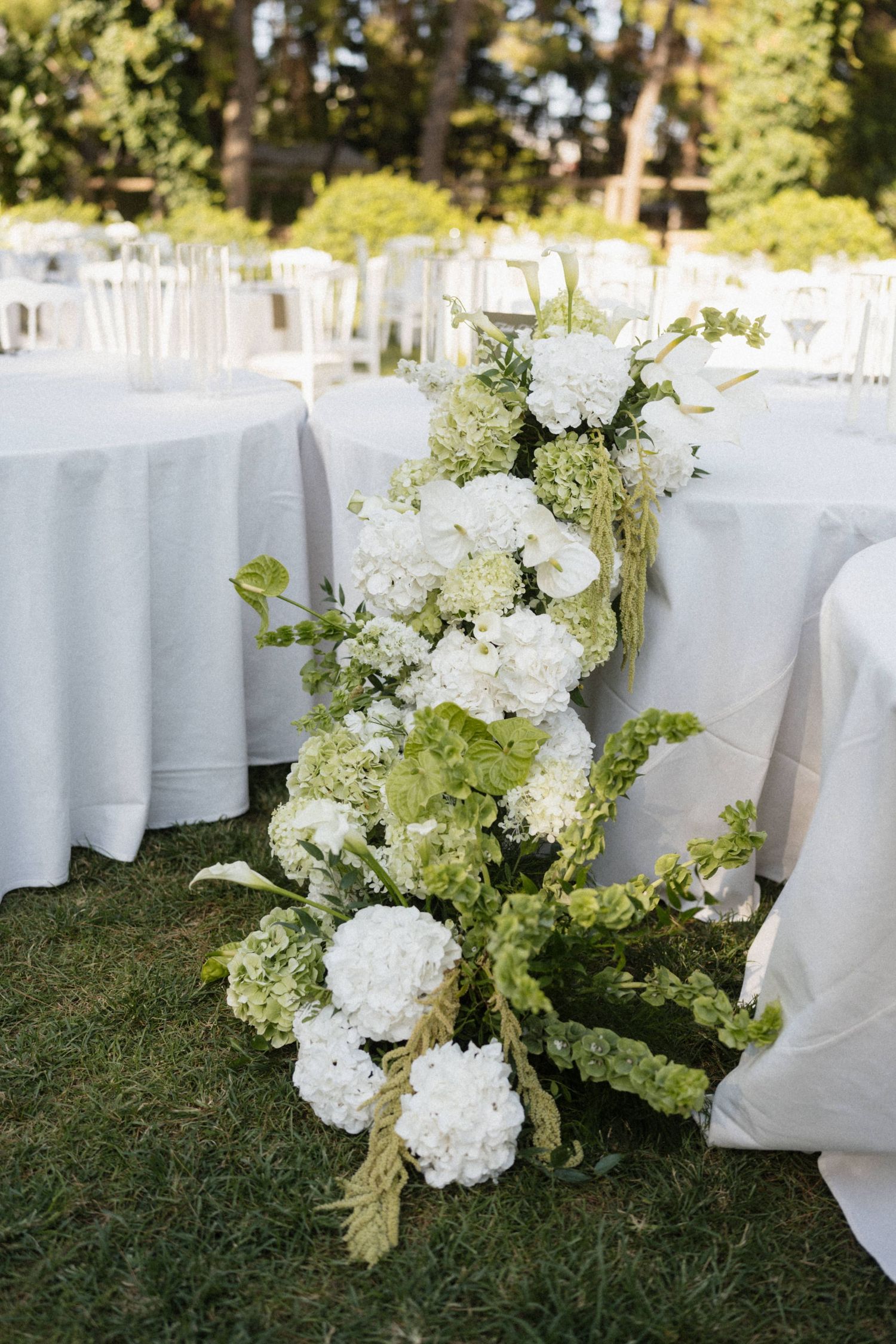 Elegant outdoor wedding reception with white and green floral arrangement on grass beside tables with white linens.