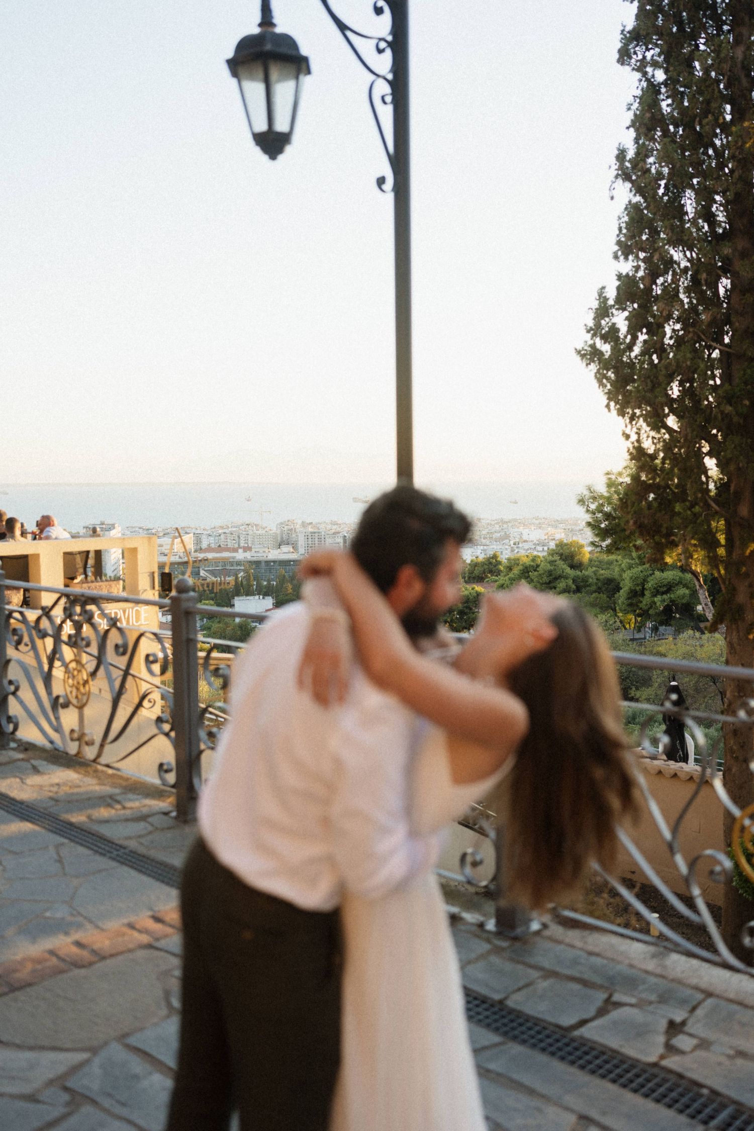 Couple sharing a romantic embrace on a scenic terrace with city views and ornate iron railings at golden hour.