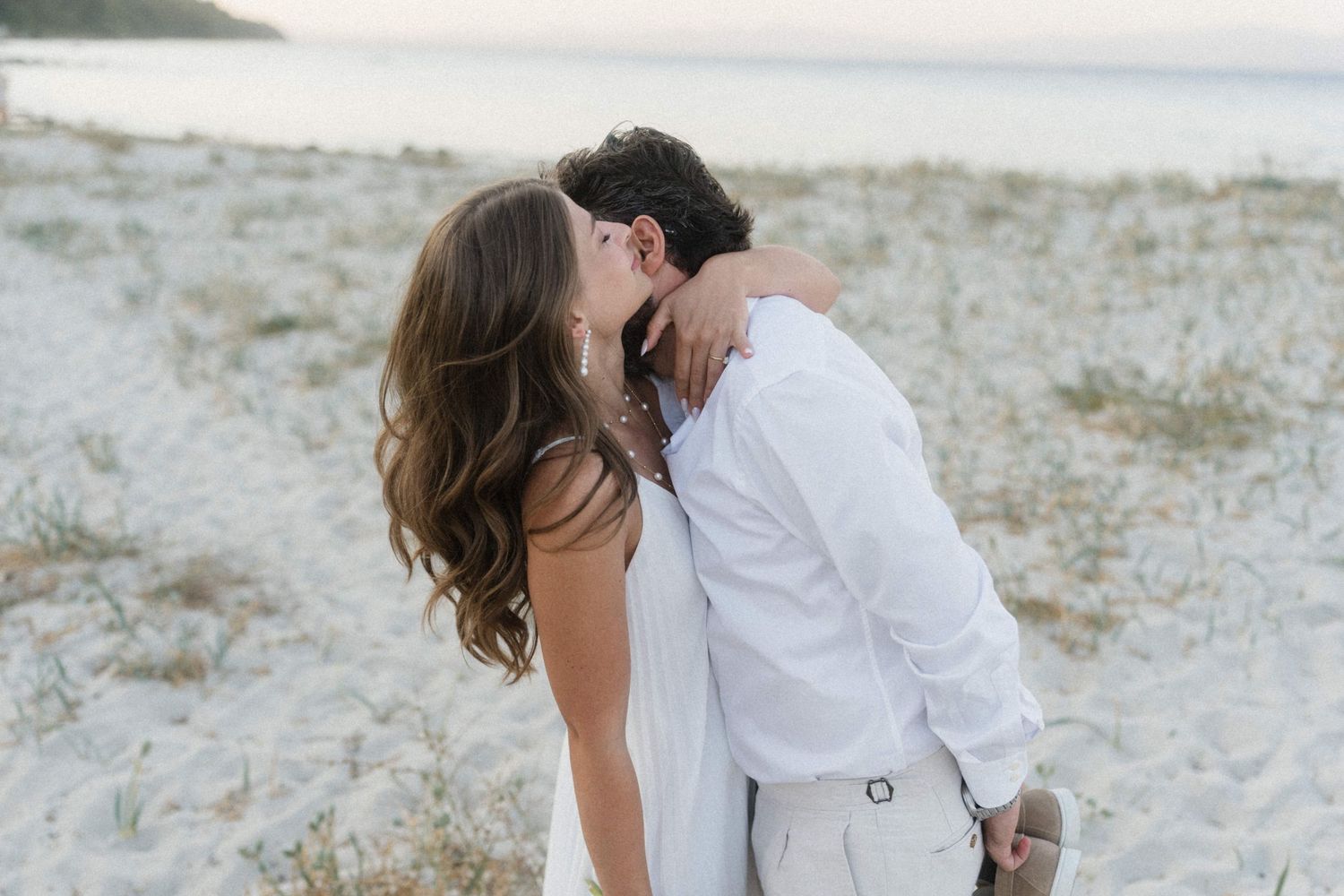 Romantic couple sharing a kiss on a white sand beach at sunset, dressed in elegant white wedding attire.