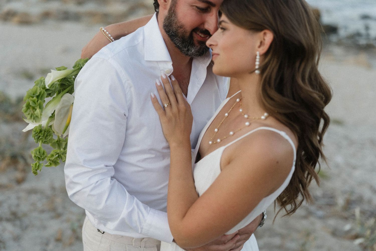 Couple in white wedding attire embracing on a beach, bride holding tropical bouquet.