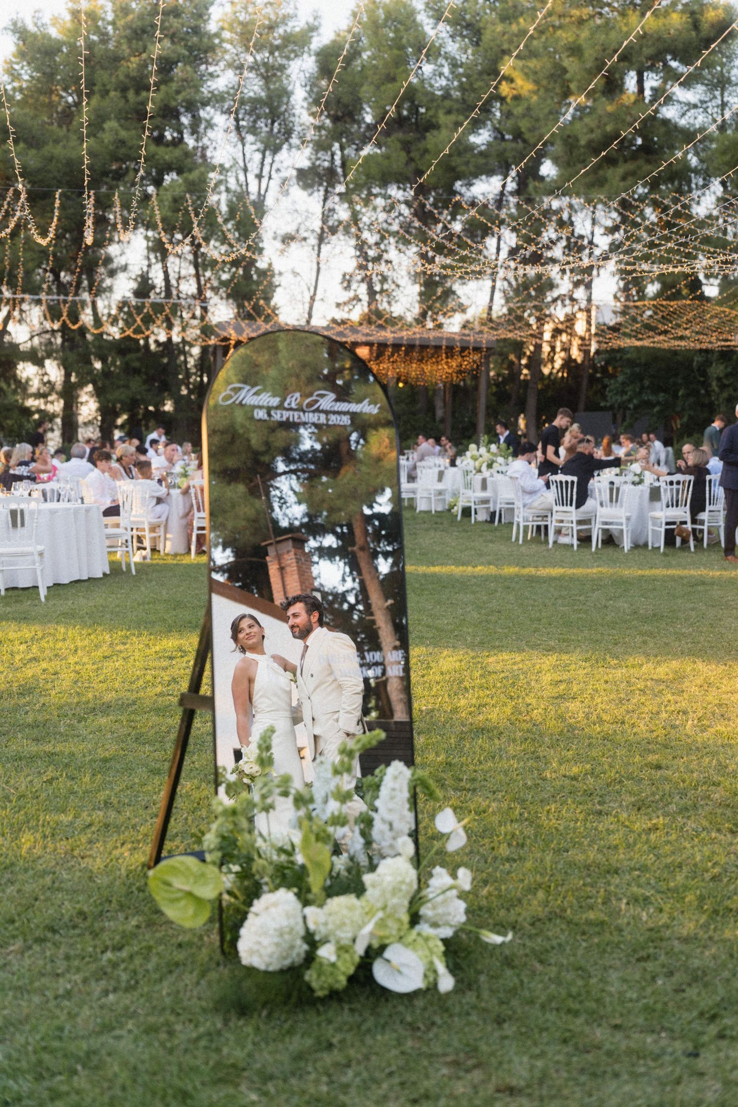 Outdoor wedding reception setup with a mirrored welcome sign, white floral arrangement, string lights, and guests seated at tables.